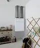 Zentrales Lüftungsgerät von Vaillant in weißer Ausführung, montiert an der Wand, mit zwei grauen Abluftrohren. Neben einem Holzgestell und einem Tisch im Raum platziert.